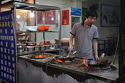 A man in a restaurant kitchen stands at a food stand with three hot wok slots, two of which are occupied by woks