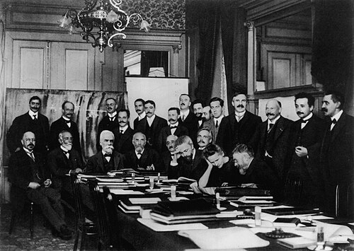 At the first Solvay Conference (1911), Curie (seated second from right) confers with Henri Poincaré. Standing nearby are Rutherford (fourth from right), Einstein (second from right), and Paul Langevin (far right).