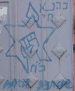 Kahanist graffiti in Hebron on a Palestinian home. The words to the top right say "Kahane Chai". The fist inside the Star of David is the party logo. Below is the acronym for "Kahane Chai" which is also the Hebrew word for strength.