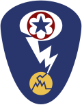 Oval shaped shoulder patch with a deep blue background. At the top is a red circle and blue star, the patch of the Army Service Forces. It is surrounded by a white oval, representing a mushroom cloud. Below it is a white lightning bolt cracking a yellow circle, representing an atom.
