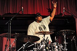 The drummer from the band Suicidal Tendencies, Eric Moore, is shown behind his drumkit. One hand is raised with the index finger and pinky extended.
