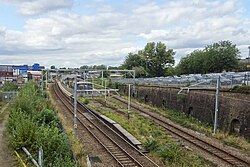 An electrified railway station which has two tracks with an island platform in the middle. The station is quite overgrown both on the platform and on the other sides of the tracks, and there is a steep cutting wall on the right-hand side