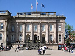 A panoramic shot of a town. In the foreground a modern-looking bus station in front of a red-brick building with large sash windows; in the background a church spire and a hill with houses on the hillside.