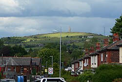 A hill in the background. The photo is taken on a street with the camera facing down the street and terraced houses on both sides. Cars, streetlights, and a 20mph speed limit sign are all visible in the foreground.