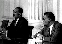 Two men in suits seated next to each other with their arms resting on a table
