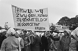 Black and white photo of a crowd, mostly women wearing headscarves, holding up a fabric sign reading: Terwijl wij zwijgen, kunnen zij gemarteld of de doodstraf krijgen