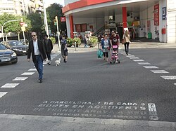 A scene of pedestrians crossing a street in Barcelona. The crossing is demarcated by white striped lines and adorned with faded, partially unreadable stenciled text in Spanish. The pedestrian signal is green, and several cars are positioned just outside the crossing.