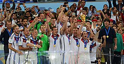 View of the celebrating German team on a podium behind a glass screen, Lahm holding the trophy aloft, while spectators with cameras stand behind