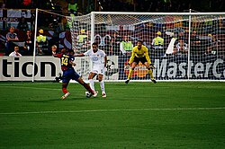 A man in a blue-and-red shirt with a football at his feet runs at a man in a white shirt. In the background, a man in a yellow shirt prepares for the ball to be kicked towards him.