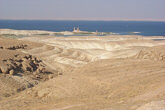 Two weathered stone towers in the background partly surrounded by the water of a river, while mounds of desert sand are pictured in the foreground