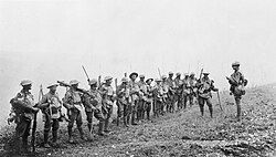  A line of soldiers in battle equipment face another soldier who is addressing them on a gentle slope.  Behind them, smoke or fog obscures the rest of the terrain.