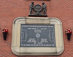 A stone tablet, inscribed with the image of a football pitch and several names. It is surrounded by a stone border in the shape of a football stadium. Above the tablet is a wooden carving of two men holding a large wreath.