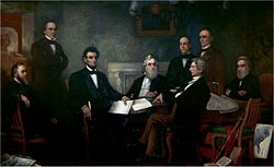 A dark-haired, bearded, middle-aged man holding documents is seated among seven other men