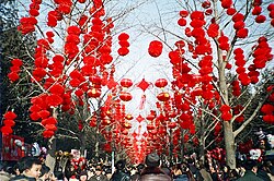 A group of people walking through an avenue over which is hung many dozens of red paper lanterns and other festive ornaments