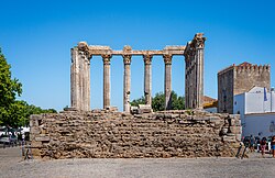 Ruins of the Roman Temple of Évora with tall Corinthian columns standing on a raised platform made of rocks in a sunlit town square, set against a bright blue sky, with a medieval tower and white buildings in the background.